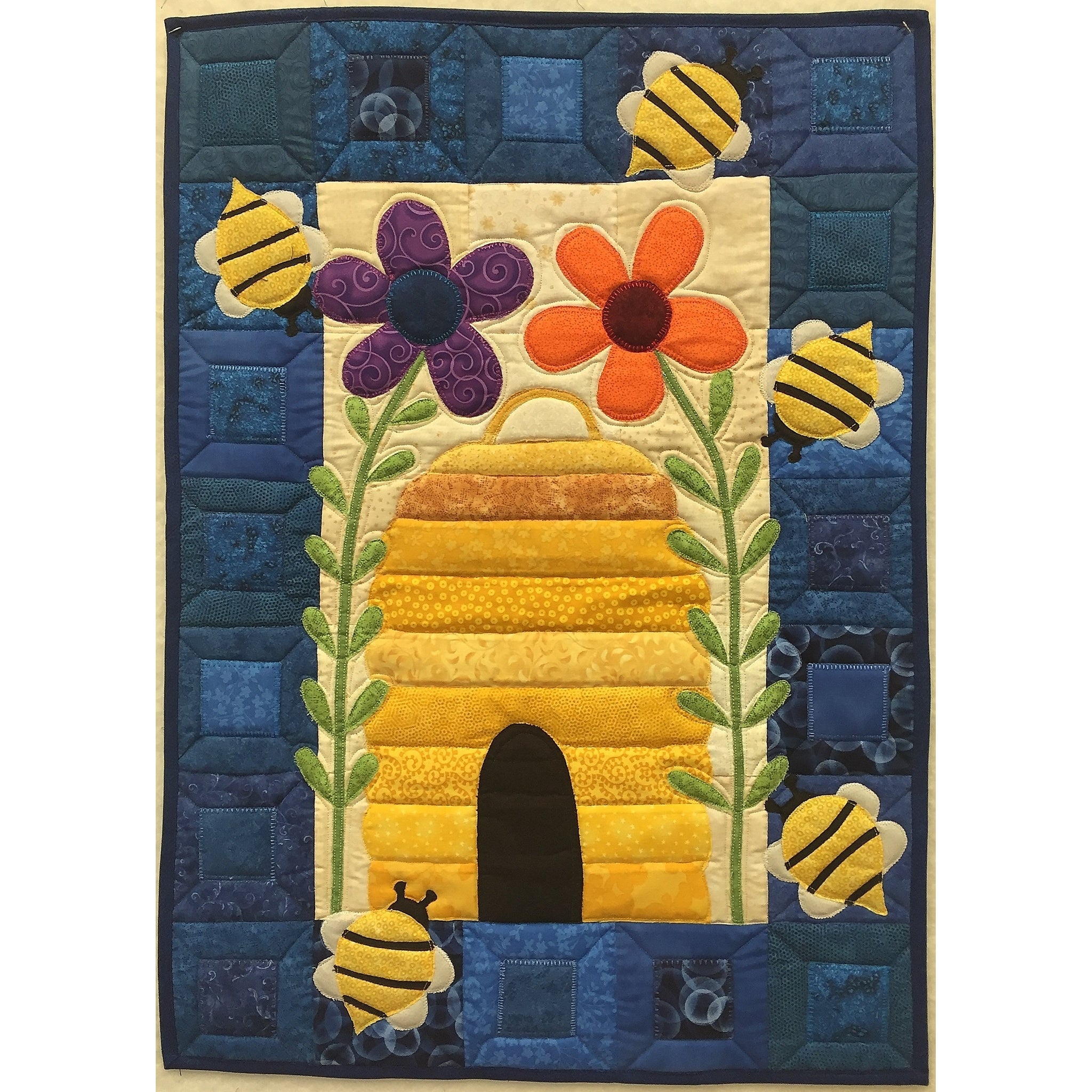A colorful quilted wall hanging featuring a yellow beehive with buzzing bees, purple and orange flowers, and a blue patchwork border.