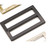 Black rectangular metal slider with double bar design, shown alongside other hardware pieces in gold and silver finishes.