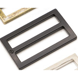 Black rectangular metal slider with double bar design, shown alongside other hardware pieces in gold and silver finishes.
