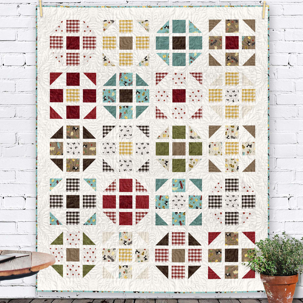 Barnyard Happy Farm quilt kit featuring geometric blocks in cream, red, yellow, teal, and brown with farm-themed prints displayed against white brick wall