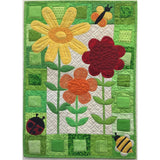 A colorful quilted wall hanging featuring three appliqué flowers in yellow, orange, and red with green stems, surrounded by a patchwork border of green fabrics and small ladybug and bee decorations.