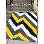 A modern chevron quilt pattern in yellow, gray, white, and black zigzag stripes displayed on concrete steps.