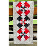 A quilted table runner featuring three pineapple blocks in red, black, and white fabrics with a cream paisley border, displayed on wooden fence rails.