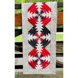 A quilted table runner featuring three pineapple blocks in red, black, and white fabrics with a cream paisley border, displayed on wooden fence rails.