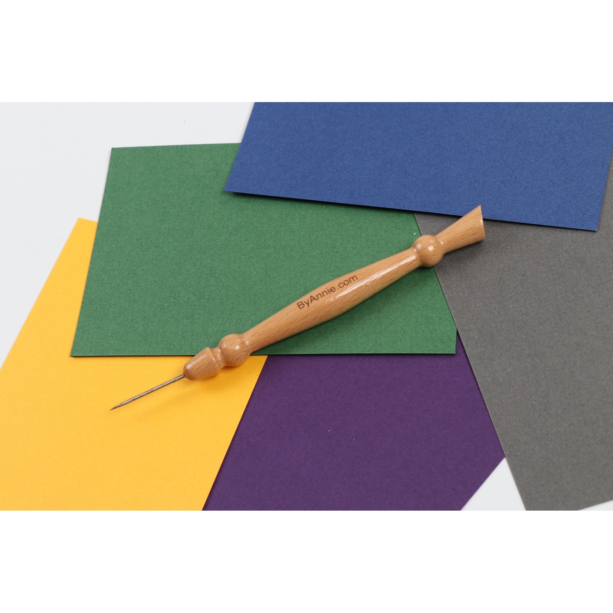 Wooden stiletto tool with pointed metal tip lying on colorful paper sheets in yellow, green, blue, purple, and gray.