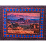 A quilted landscape artwork featuring red rock formations and desert scenery, framed by a decorative border of blue diamonds on red triangular backgrounds with purple outer edging.