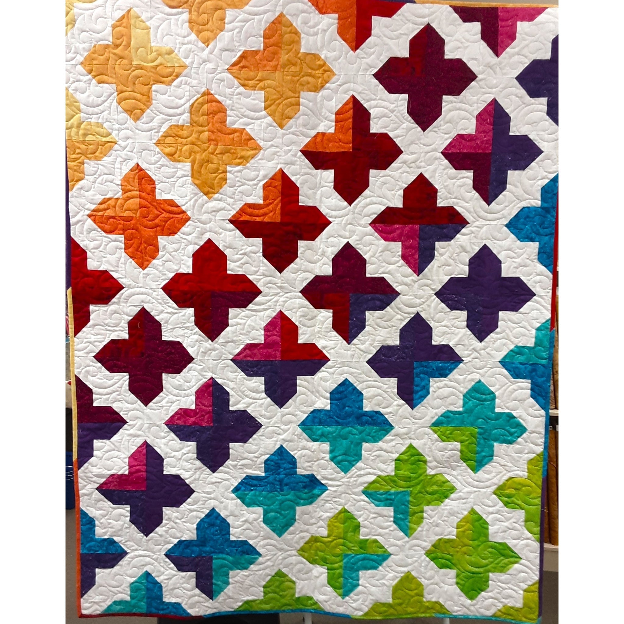 Colorful geometric quilt featuring cross-shaped blocks in a rainbow gradient from warm oranges and reds to cool blues and greens on white background.